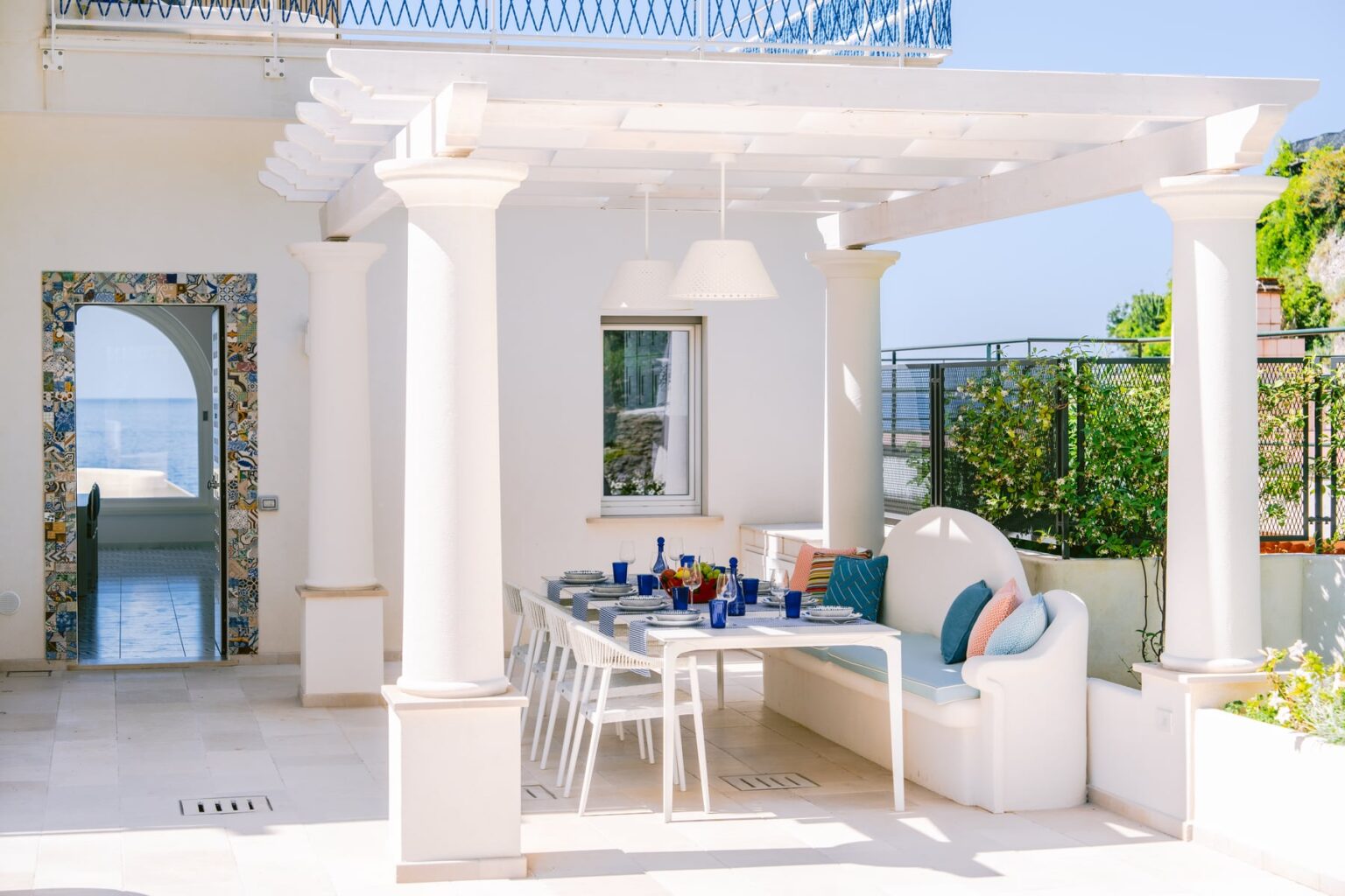 Elegant outdoor dining area featuring a white pergola, a modern table set with blue glassware, and comfortable seating, surrounded by lush greenery and a scenic view. Perfect for alfresco dining and relaxation. | Only on SopranoVillas