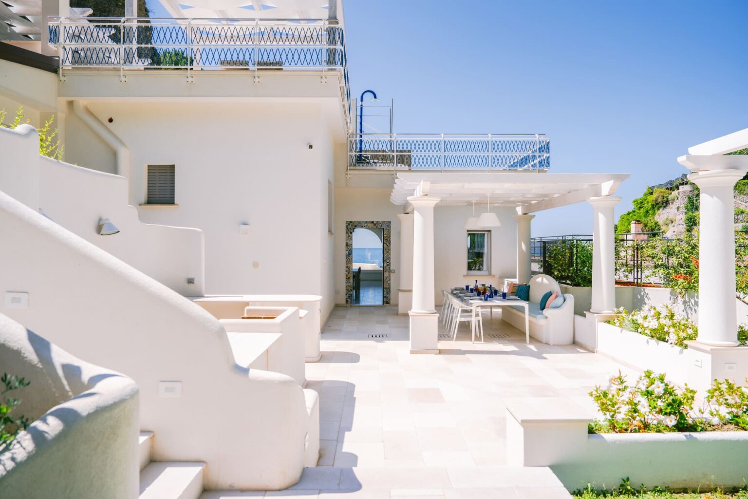 Elegant outdoor terrace of a modern villa featuring white stone architecture, a dining area, and scenic views of the ocean under a clear blue sky. | Only on SopranoVillas