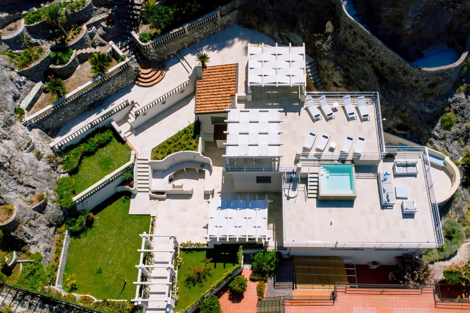 Aerial view of a luxurious villa featuring a spacious terrace with sun loungers, a private jacuzzi, landscaped gardens, and stone pathways, surrounded by rocky cliffs and greenery. Ideal for relaxation and outdoor gatherings. | Only on SopranoVillas