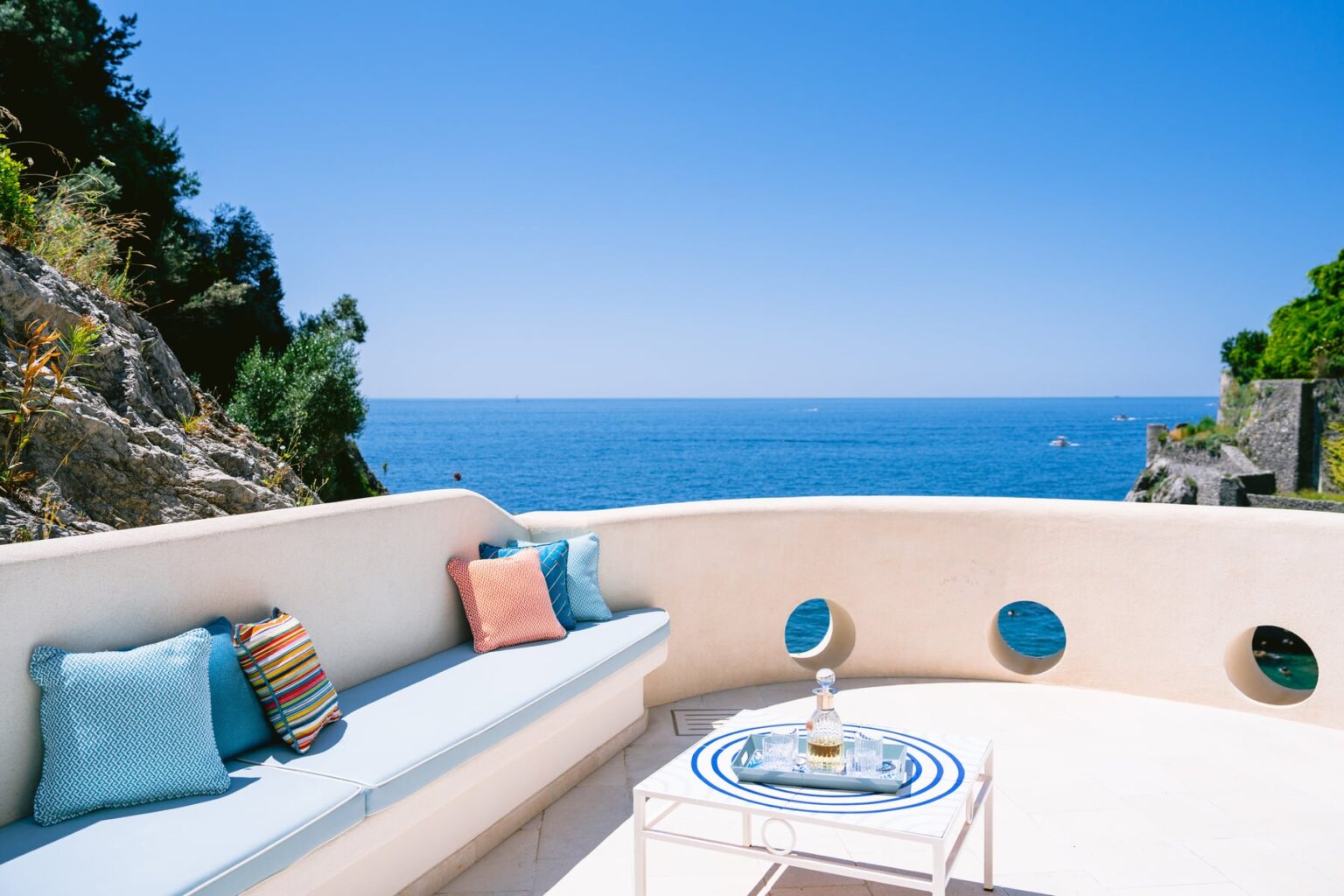 Cozy outdoor seating area with colorful pillows, overlooking a serene ocean view under a clear blue sky. Ideal for relaxation and enjoying coastal scenery. | Only on SopranoVillas