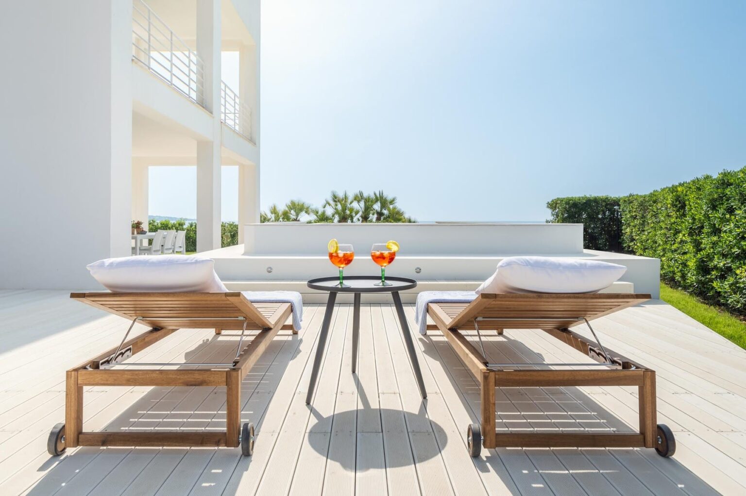 Modern outdoor patio featuring two wooden lounge chairs and a small table with refreshing drinks, surrounded by lush greenery and a clear blue sky. Ideal for relaxation and outdoor entertaining. | Only on SopranoVillas