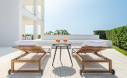 Modern outdoor patio featuring two wooden lounge chairs and a small table with refreshing drinks, surrounded by lush greenery and a clear blue sky. Ideal for relaxation and outdoor entertaining.