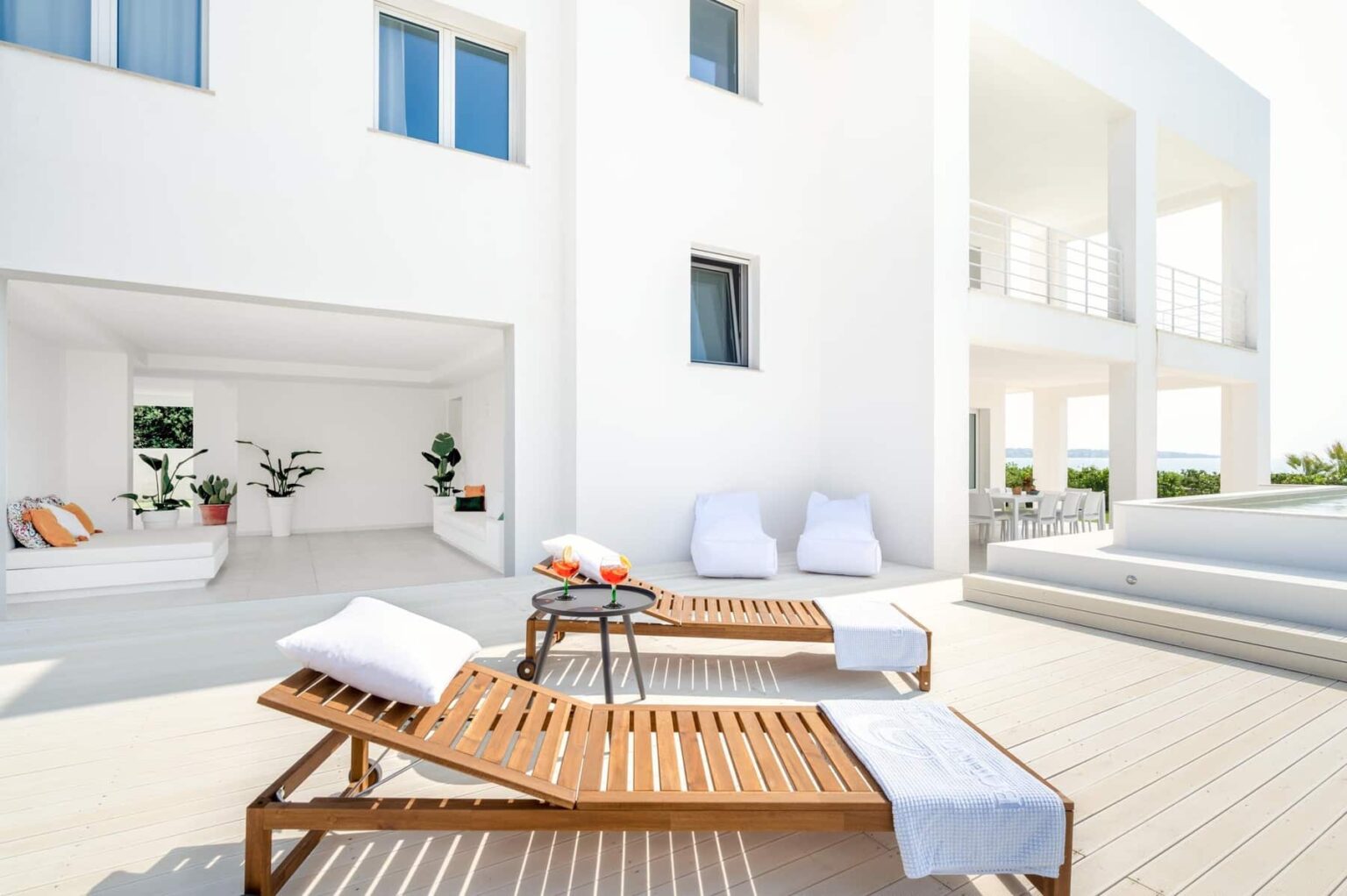 Modern white villa with a spacious outdoor terrace featuring wooden lounge chairs, white towels, and a relaxing ambiance. Lush greenery and minimalist design elements enhance the serene atmosphere. | Only on SopranoVillas