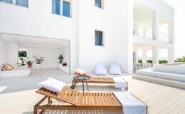 Modern white villa with a spacious outdoor terrace featuring wooden lounge chairs, white towels, and a relaxing ambiance. Lush greenery and minimalist design elements enhance the serene atmosphere.