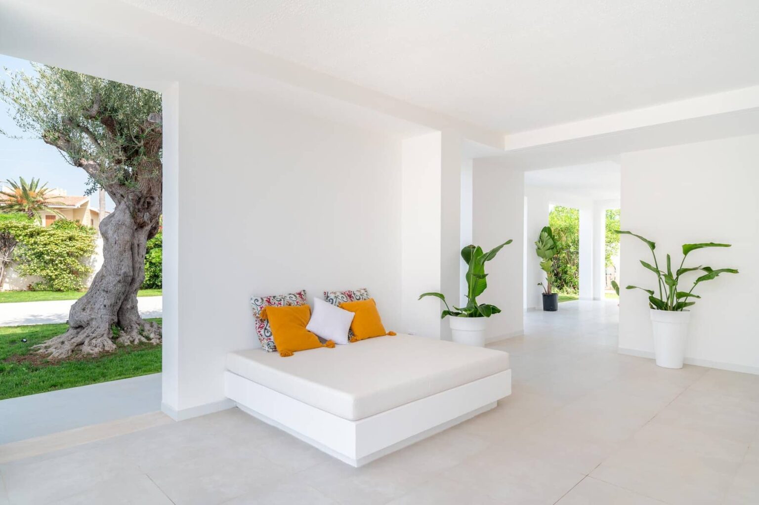 Bright and airy interior of a modern living space featuring a white sofa with orange cushions, surrounded by lush greenery and large windows allowing natural light to fill the room. | Only on SopranoVillas