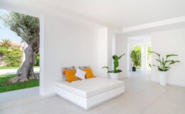 Bright and airy interior of a modern living space featuring a white sofa with orange cushions, surrounded by lush greenery and large windows allowing natural light to fill the room.