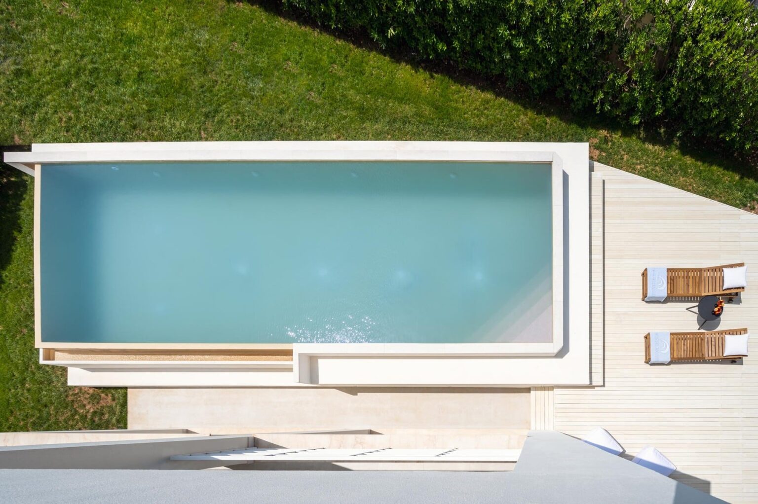 Aerial view of a modern swimming pool surrounded by lush green grass, featuring wooden lounge chairs and a stylish deck for relaxation. | Only on SopranoVillas