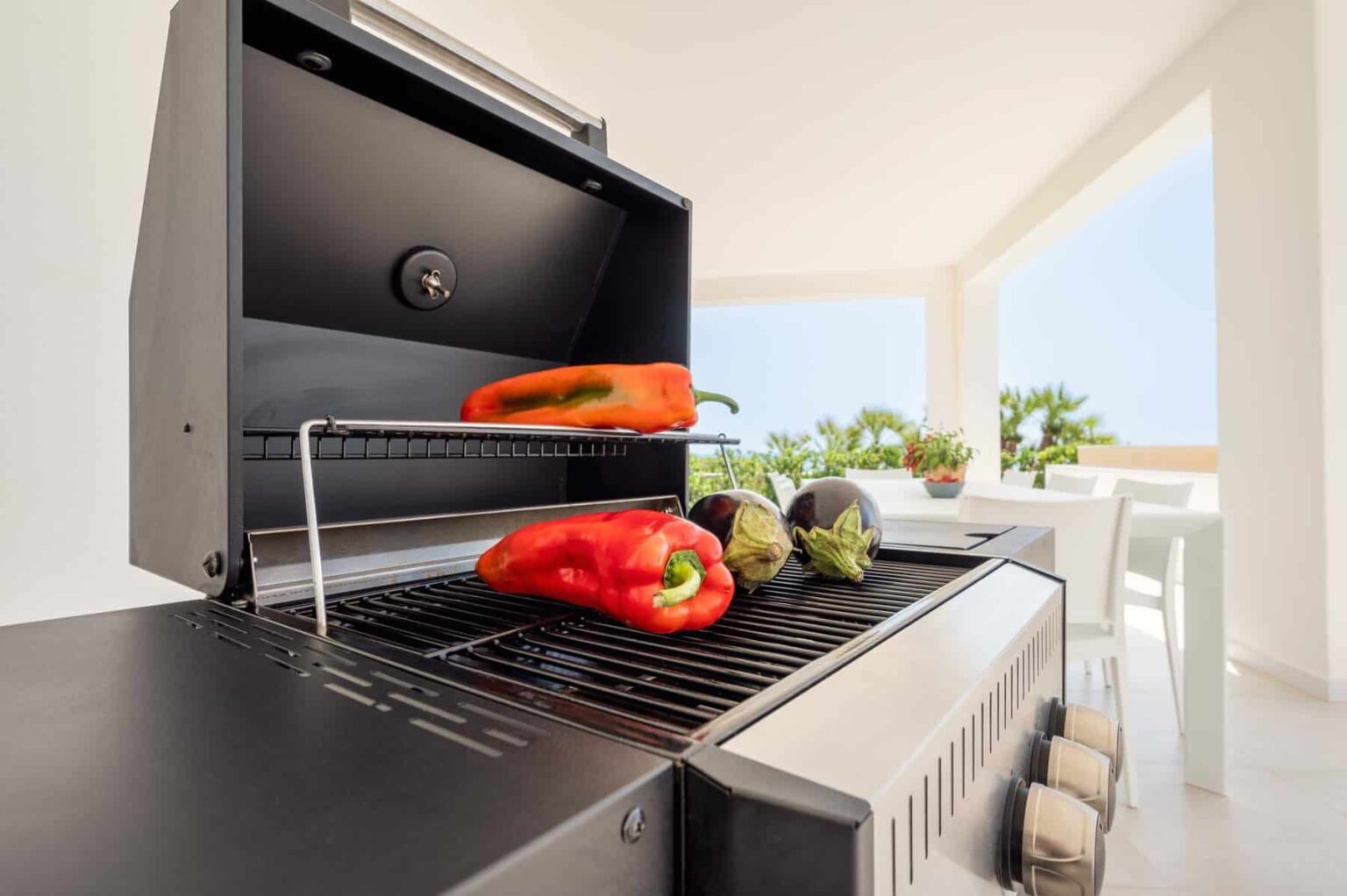 Grill with fresh red bell peppers and eggplants on the rack, set in a bright, modern outdoor kitchen with a view of greenery. Perfect for summer cooking and healthy meals. | Only on SopranoVillas