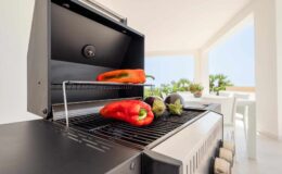 Grill with fresh red bell peppers and eggplants on the rack, set in a bright, modern outdoor kitchen with a view of greenery. Perfect for summer cooking and healthy meals.