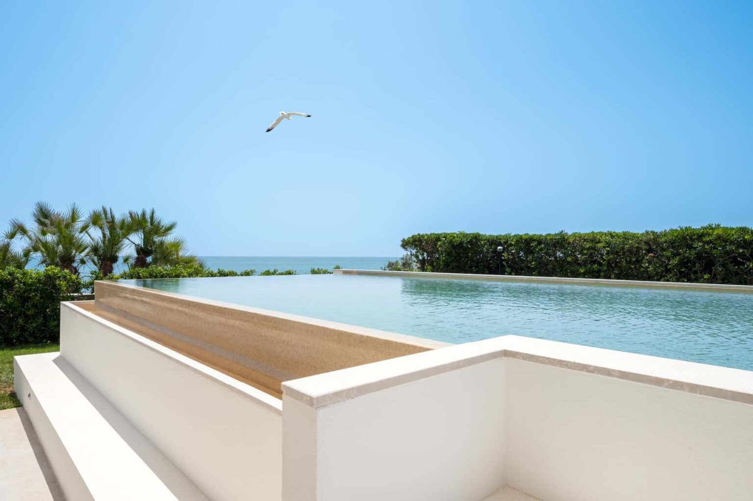Modern beachfront infinity pool overlooking the ocean with clear blue skies and lush greenery in the background. | Only on SopranoVillas