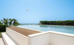 Modern beachfront infinity pool overlooking the ocean with clear blue skies and lush greenery in the background.