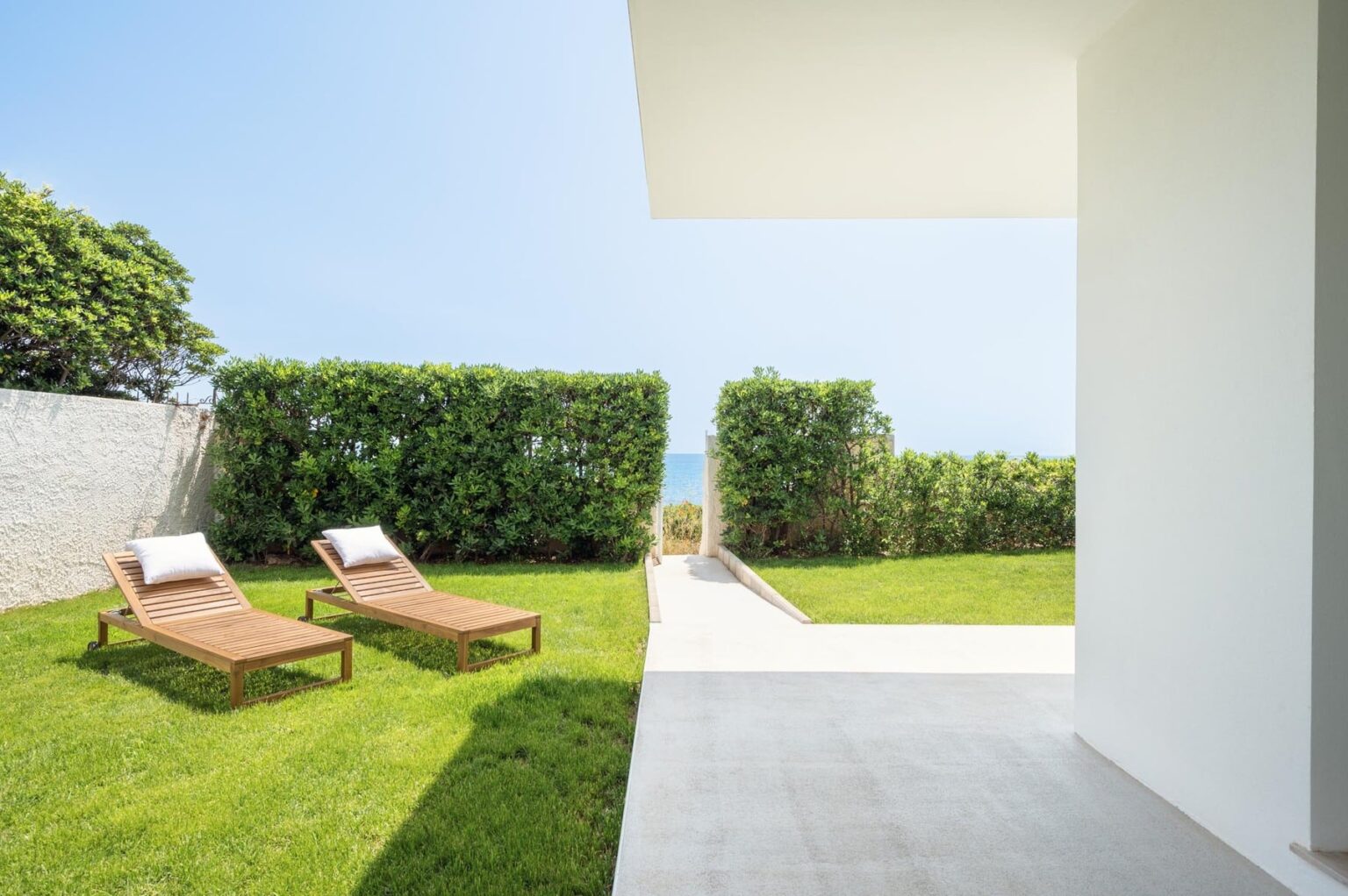 Modern outdoor patio with two lounge chairs on a manicured lawn, surrounded by lush greenery and a clear blue sky, leading to a scenic view. Ideal for relaxation and outdoor living. | Only on SopranoVillas