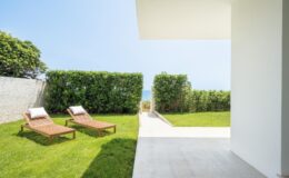 Modern outdoor patio with two lounge chairs on a manicured lawn, surrounded by lush greenery and a clear blue sky, leading to a scenic view. Ideal for relaxation and outdoor living.