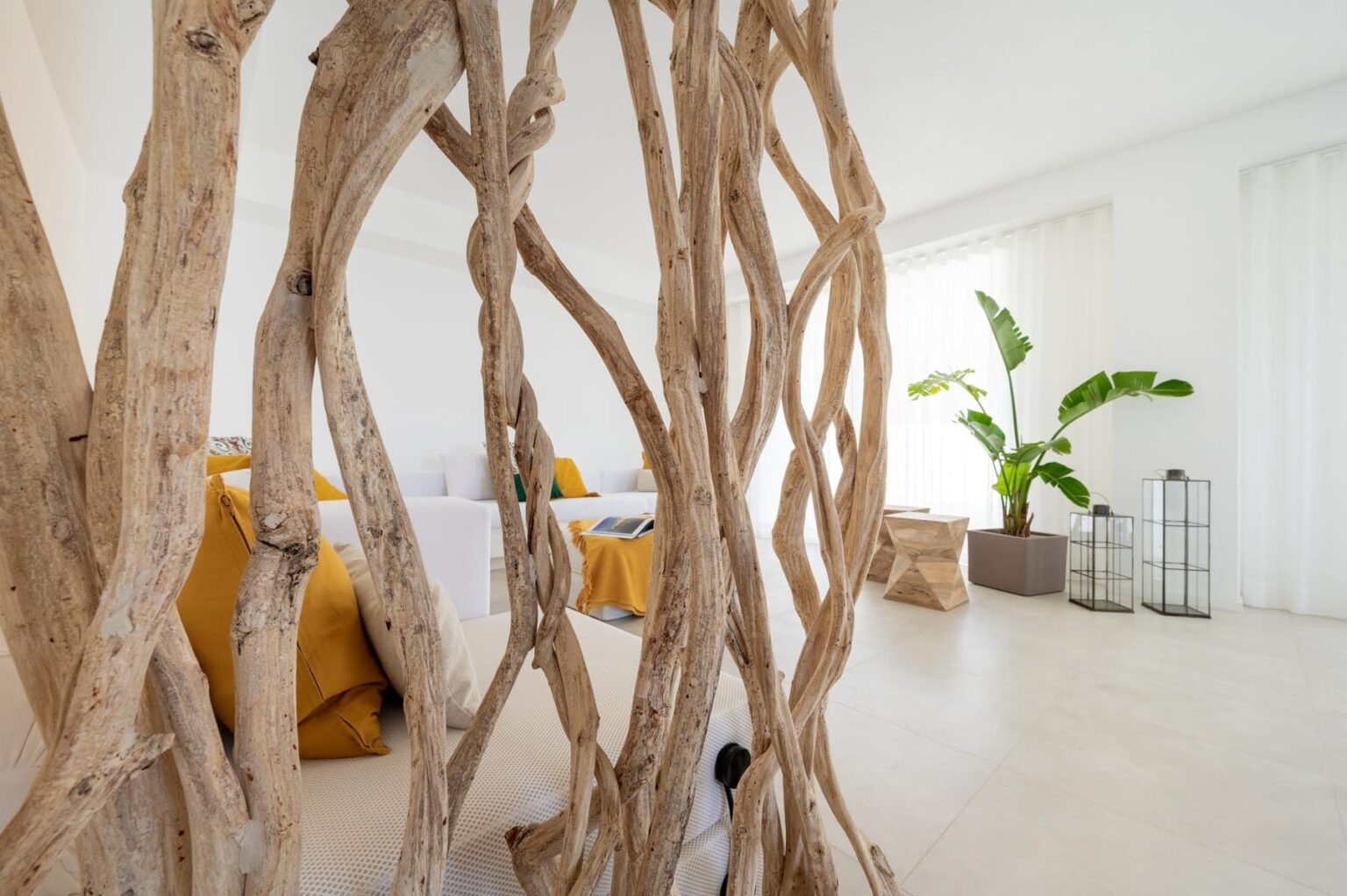 Interior view of a modern living space featuring a decorative wooden partition, yellow cushions, and a green plant. The room has bright natural light streaming through sheer curtains, highlighting the minimalist design and contemporary furniture. | Only on SopranoVillas