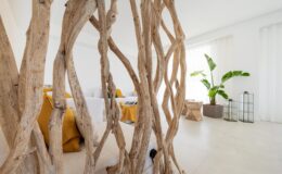 Interior view of a modern living space featuring a decorative wooden partition, yellow cushions, and a green plant. The room has bright natural light streaming through sheer curtains, highlighting the minimalist design and contemporary furniture.