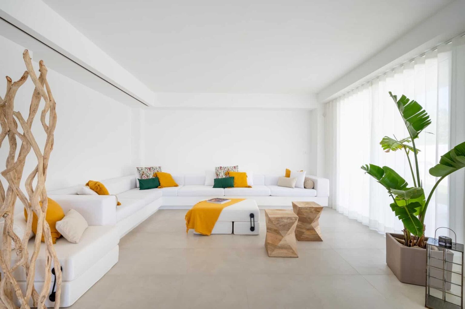 Modern living room featuring a white sectional sofa adorned with colorful throw pillows, a minimalist wooden coffee table, and large windows allowing natural light. Lush green plants add a touch of nature to the contemporary decor. | Only on SopranoVillas