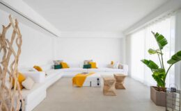 Modern living room featuring a white sectional sofa adorned with colorful throw pillows, a minimalist wooden coffee table, and large windows allowing natural light. Lush green plants add a touch of nature to the contemporary decor.