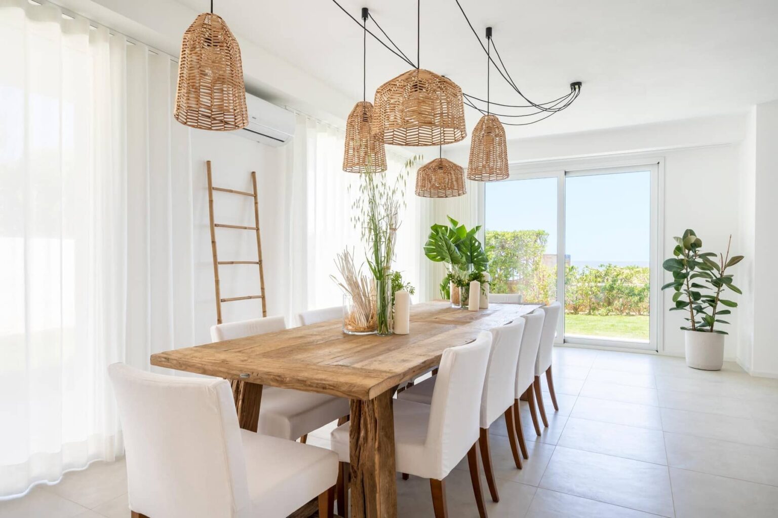 Modern dining room featuring a large wooden table surrounded by white chairs, adorned with natural woven pendant lights. Bright, airy space with large windows and greenery, creating a fresh and inviting atmosphere. | Only on SopranoVillas