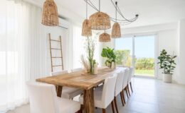 Modern dining room featuring a large wooden table surrounded by white chairs, adorned with natural woven pendant lights. Bright, airy space with large windows and greenery, creating a fresh and inviting atmosphere.