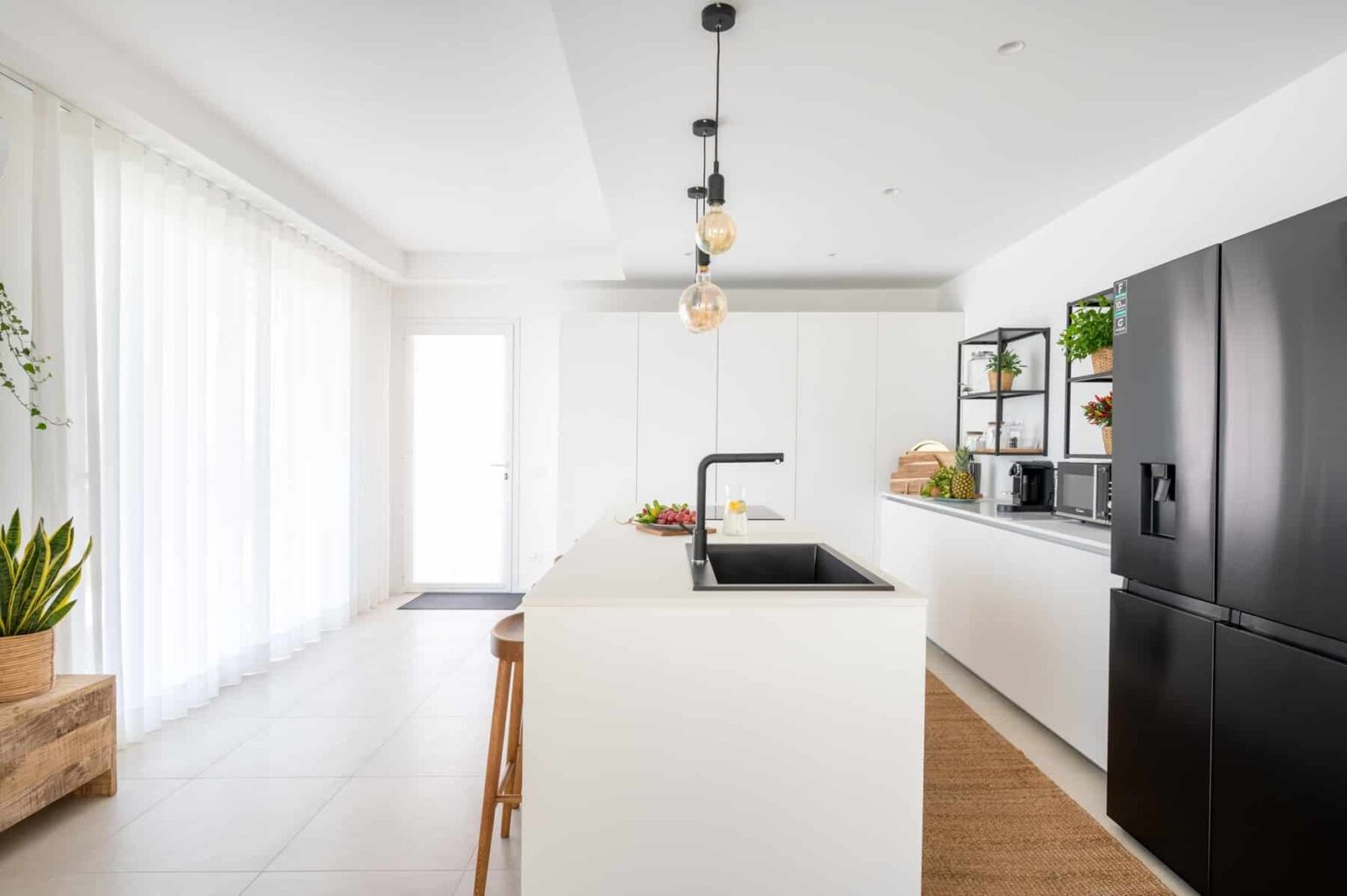 Modern kitchen featuring a sleek white design, black accents, and natural light. Includes a central island with a black sink, wooden bar stool, and fresh fruit display. The kitchen is equipped with minimalist cabinetry, a black refrigerator, and open shelving with plants and kitchen appliances. | Only on SopranoVillas