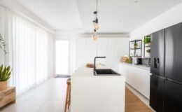 Modern kitchen featuring a sleek white design, black accents, and natural light. Includes a central island with a black sink, wooden bar stool, and fresh fruit display. The kitchen is equipped with minimalist cabinetry, a black refrigerator, and open shelving with plants and kitchen appliances.