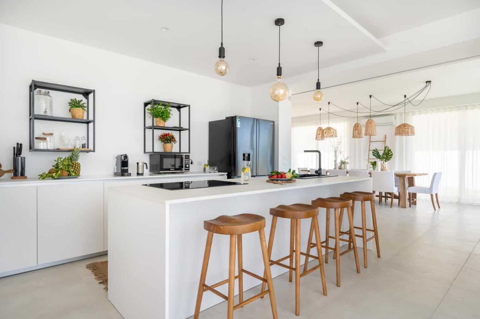 Modern kitchen interior featuring a spacious island with wooden bar stools, sleek white cabinetry, and contemporary lighting. The design includes open shelving with plants and kitchen appliances, creating a bright and inviting atmosphere. | Only on SopranoVillas