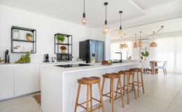 Modern kitchen interior featuring a spacious island with wooden bar stools, sleek white cabinetry, and contemporary lighting. The design includes open shelving with plants and kitchen appliances, creating a bright and inviting atmosphere.