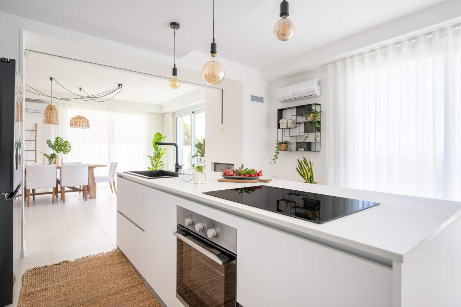 Modern white kitchen with an island, featuring a built-in cooktop, oven, and sleek cabinetry. Natural light filters through large windows, illuminating the space adorned with indoor plants and stylish pendant lighting. | Only on SopranoVillas