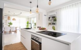 Modern white kitchen with an island, featuring a built-in cooktop, oven, and sleek cabinetry. Natural light filters through large windows, illuminating the space adorned with indoor plants and stylish pendant lighting.