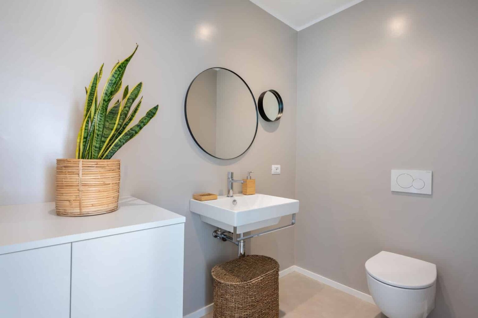Modern bathroom interior featuring a sleek sink, round mirror, and minimalistic decor. A potted plant adds a touch of greenery, while neutral walls and stylish storage enhance the contemporary design. Ideal for home improvement inspiration. | Only on SopranoVillas