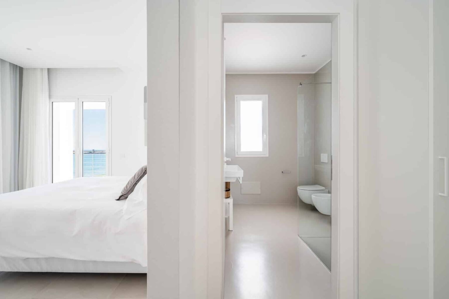 Modern bedroom with a white bed and large windows offering a view of the ocean, leading into a minimalist bathroom featuring a glass shower and sleek fixtures. Bright, airy design emphasizes tranquility and comfort. | Only on SopranoVillas