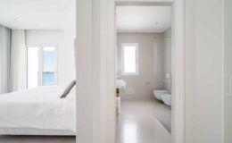 Modern bedroom with a white bed and large windows offering a view of the ocean, leading into a minimalist bathroom featuring a glass shower and sleek fixtures. Bright, airy design emphasizes tranquility and comfort.