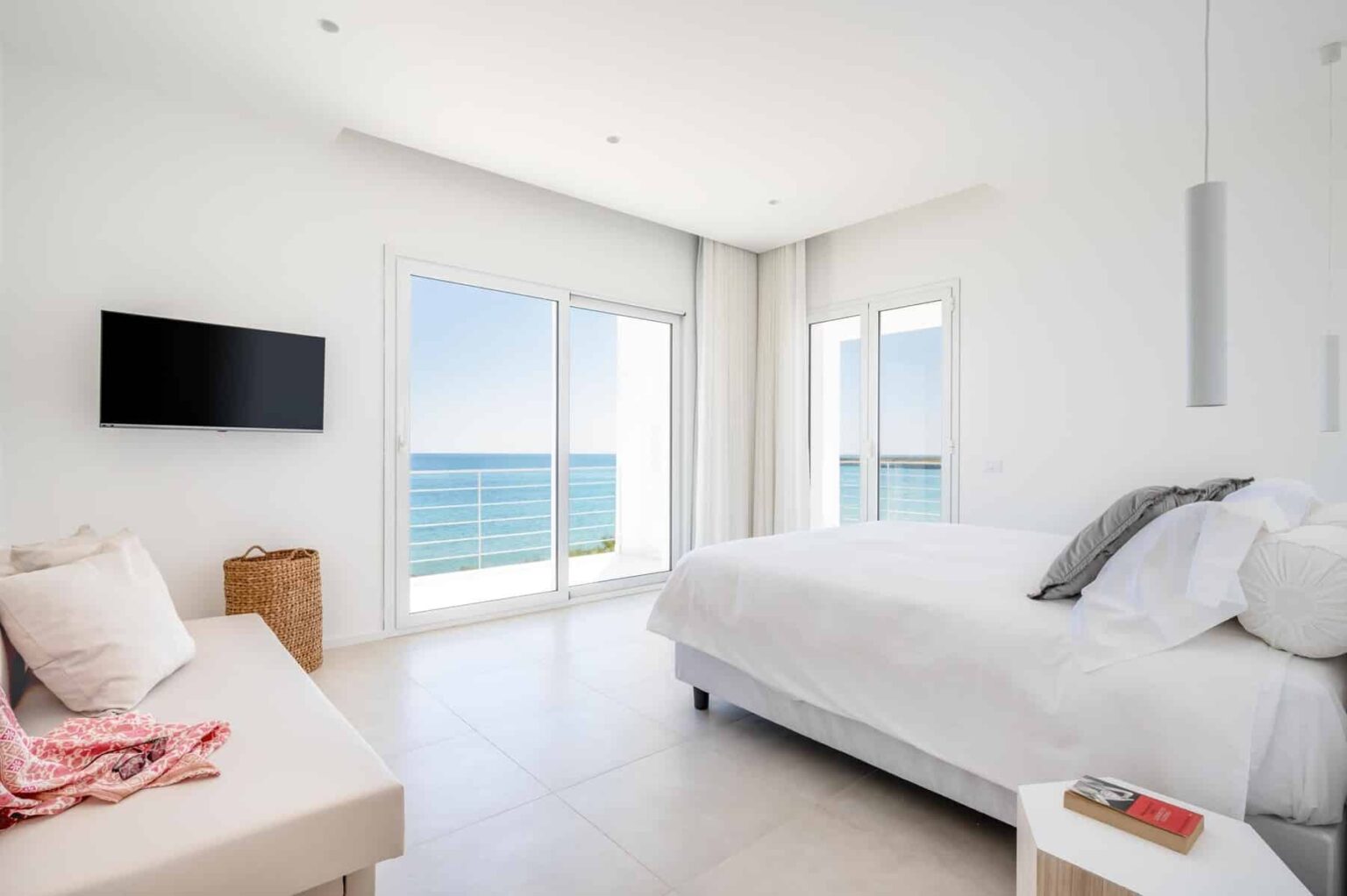 Modern coastal bedroom with a large window overlooking the sea, featuring a white bed, minimalist decor, and a wall-mounted TV. | Only on SopranoVillas