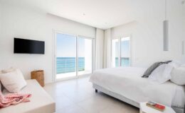 Modern coastal bedroom with a large window overlooking the sea, featuring a white bed, minimalist decor, and a wall-mounted TV.