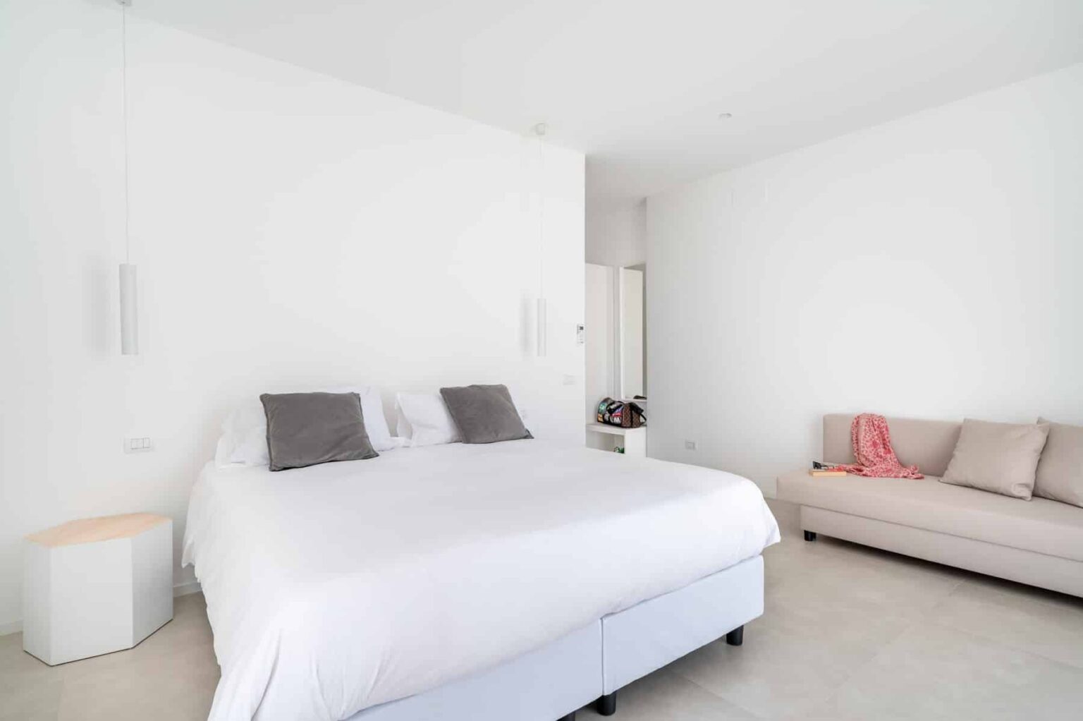 Modern bedroom featuring a large white bed with gray pillows, minimalistic decor, and a comfortable beige sofa against a bright white wall. Ideal for showcasing contemporary interior design. | Only on SopranoVillas