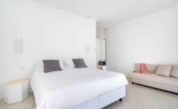 Modern bedroom featuring a large white bed with gray pillows, minimalistic decor, and a comfortable beige sofa against a bright white wall. Ideal for showcasing contemporary interior design.