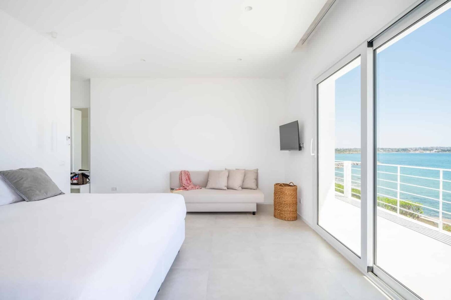 Modern beachfront bedroom with a large window showcasing ocean views, featuring a white bed, a cozy sofa, and minimalist decor. | Only on SopranoVillas