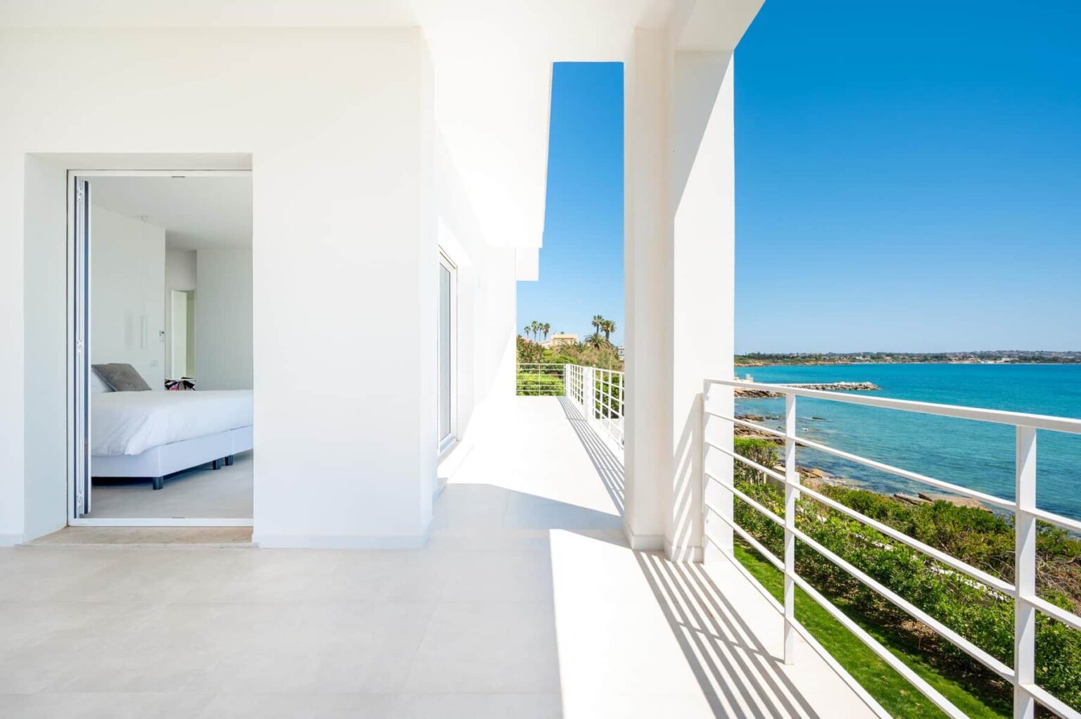 Modern balcony overlooking a serene ocean view with a bright, sunlit interior featuring a white bedroom. Ideal for luxury vacation rentals or real estate listings. | Only on SopranoVillas