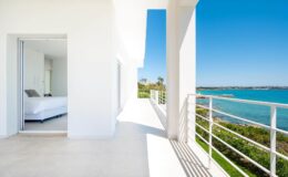Modern balcony overlooking a serene ocean view with a bright, sunlit interior featuring a white bedroom. Ideal for luxury vacation rentals or real estate listings.