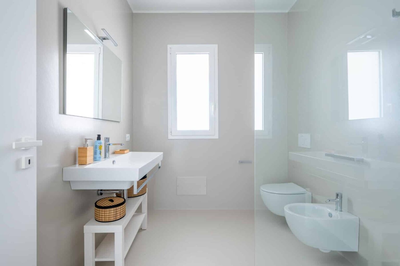 Modern bathroom design featuring a minimalist aesthetic, white walls, natural light from windows, a sleek vanity with toiletries, and stylish storage baskets. | Only on SopranoVillas