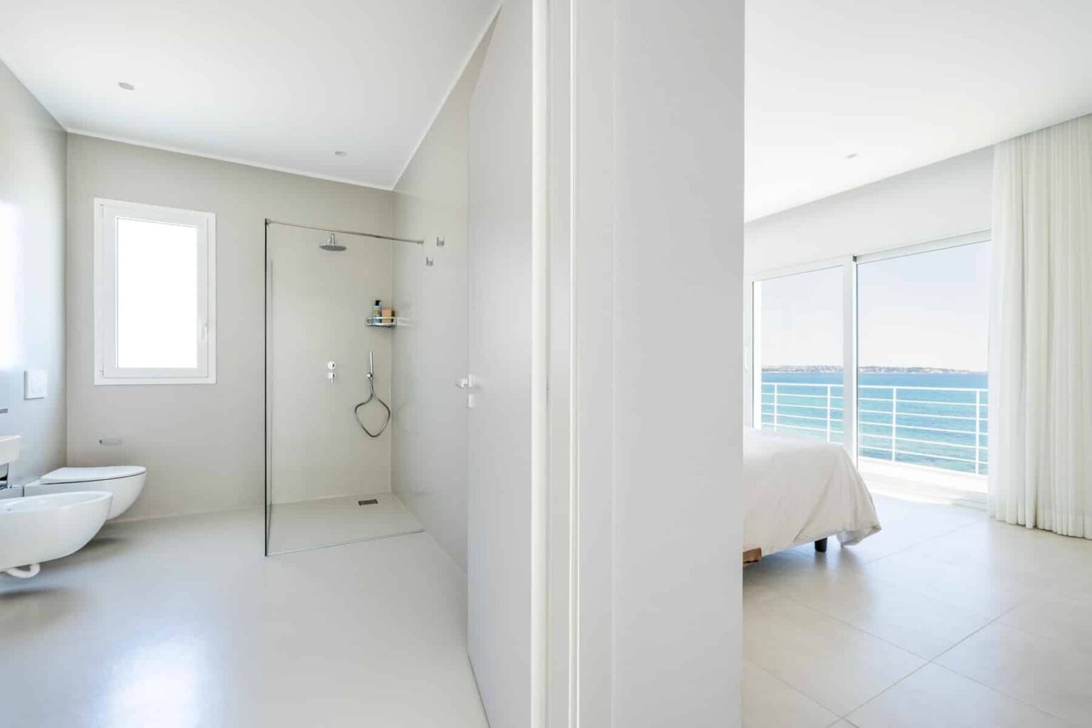 Modern bathroom and bedroom interior with large windows offering ocean views, featuring a glass shower and minimalistic design elements. | Only on SopranoVillas
