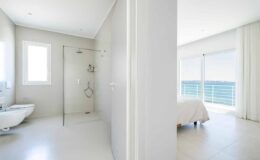 Modern bathroom and bedroom interior with large windows offering ocean views, featuring a glass shower and minimalistic design elements.