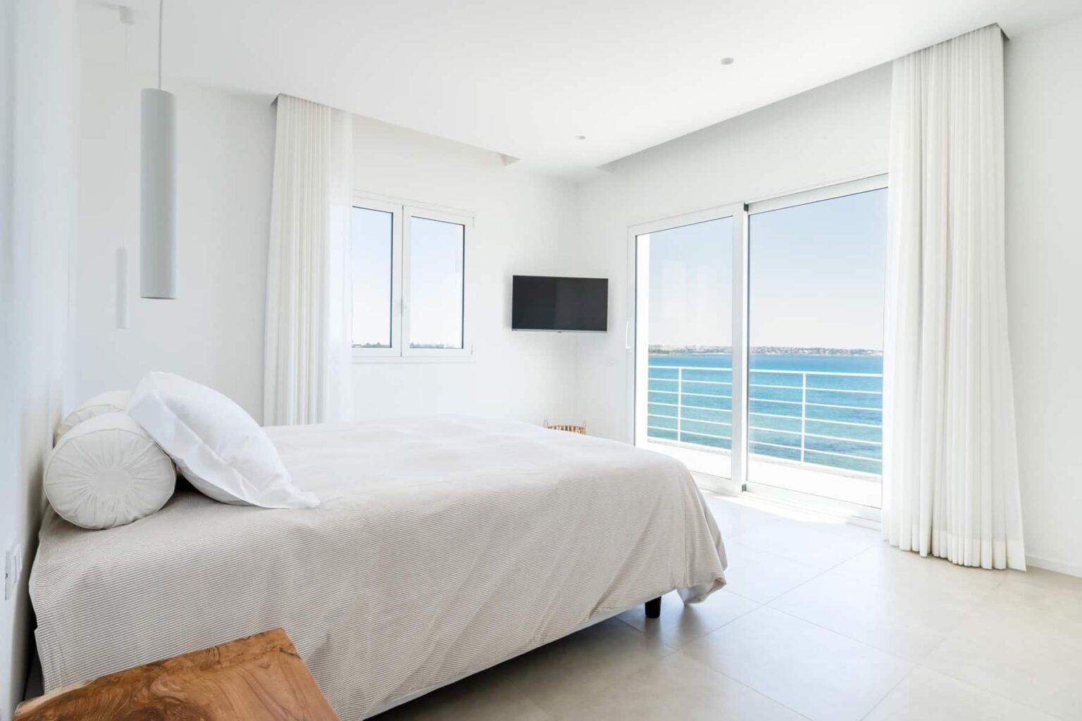 Modern bedroom with a large window and balcony overlooking the ocean, featuring a comfortable bed with light bedding, minimalist decor, and a wall-mounted TV. | Only on SopranoVillas