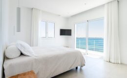 Modern bedroom with a large window and balcony overlooking the ocean, featuring a comfortable bed with light bedding, minimalist decor, and a wall-mounted TV.