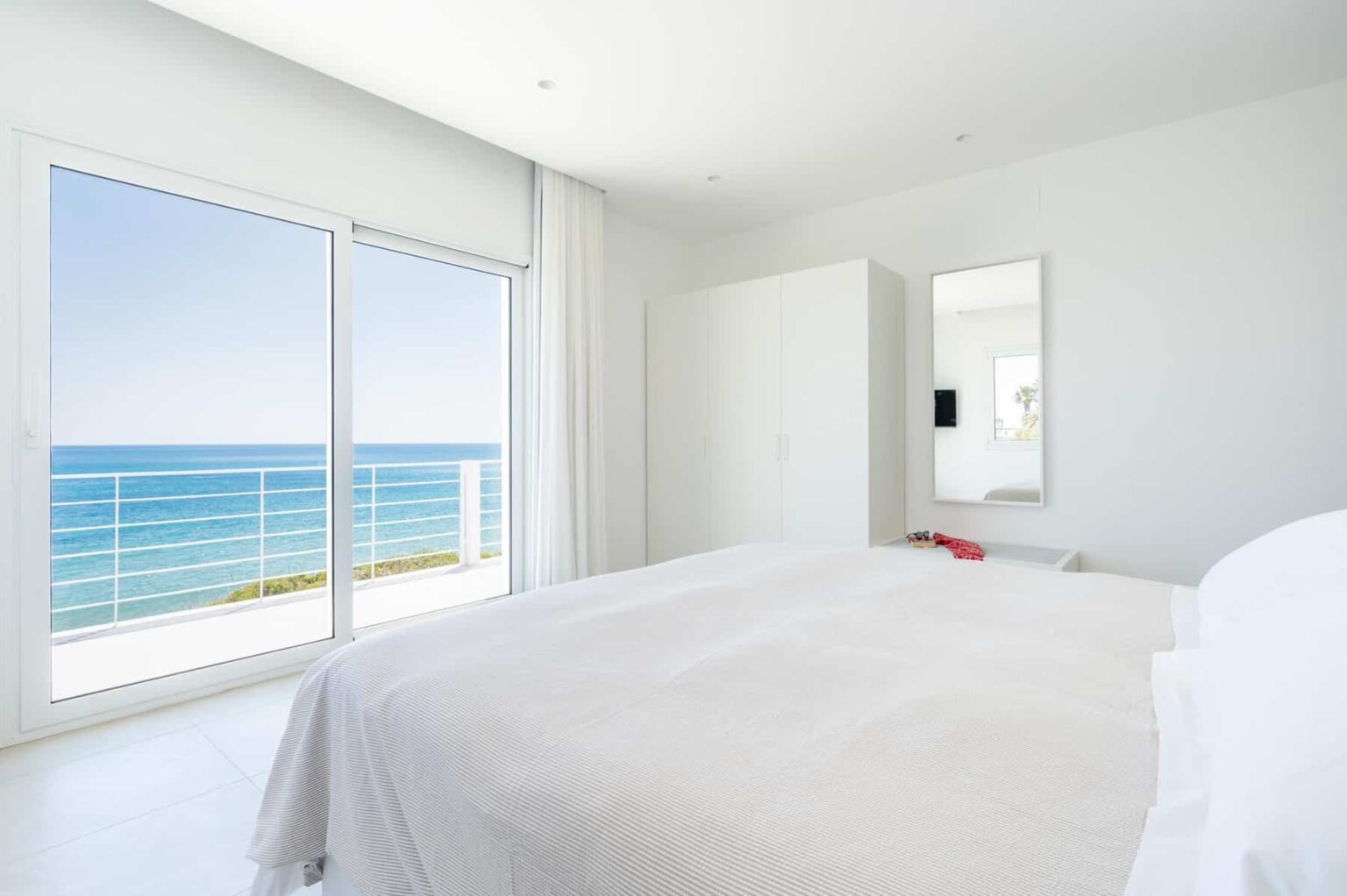 Bright and airy bedroom with a white bedspread, featuring large windows that offer a stunning view of the ocean. The room has minimalist decor, including a wardrobe and a mirror, creating a serene coastal retreat. | Only on SopranoVillas
