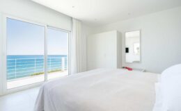 Bright and airy bedroom with a white bedspread, featuring large windows that offer a stunning view of the ocean. The room has minimalist decor, including a wardrobe and a mirror, creating a serene coastal retreat.