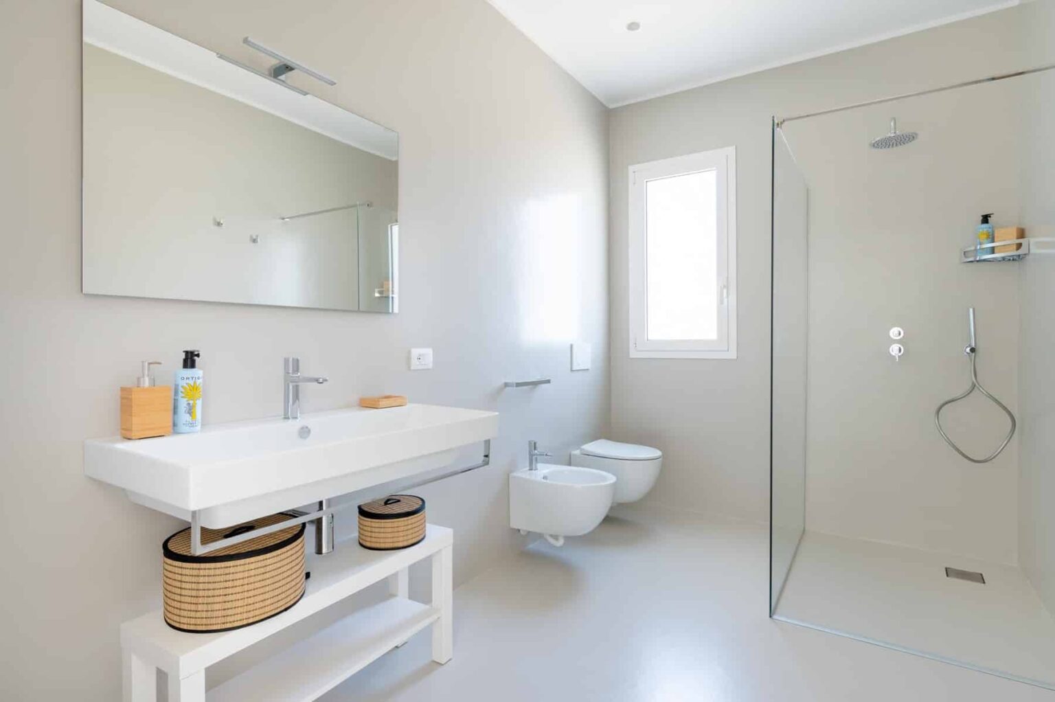 Modern bathroom design featuring a sleek white vanity with a large mirror, minimalist shower enclosure, and natural light from a window. The space includes a contemporary toilet and stylish storage baskets, showcasing a clean and functional aesthetic. | Only on SopranoVillas
