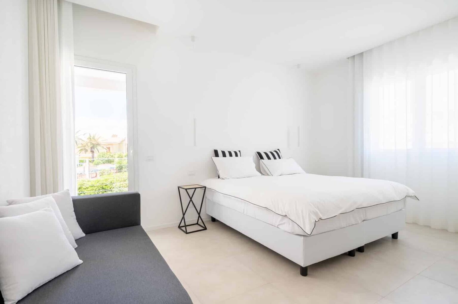 Modern bedroom with a queen-sized bed featuring striped pillows, a gray sofa with white cushions, and large windows allowing natural light. Minimalist decor emphasizes a bright and airy atmosphere. | Only on SopranoVillas