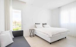 Modern bedroom with a queen-sized bed featuring striped pillows, a gray sofa with white cushions, and large windows allowing natural light. Minimalist decor emphasizes a bright and airy atmosphere.