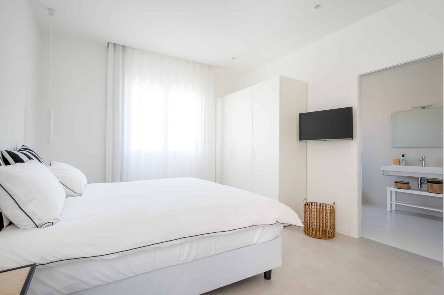 Bright and modern bedroom featuring a queen-sized bed with white linens, a flat-screen TV mounted on the wall, and a spacious wardrobe. Natural light filters through sheer curtains, creating a serene atmosphere. A glimpse of a stylish bathroom is visible through an open doorway. | Only on SopranoVillas